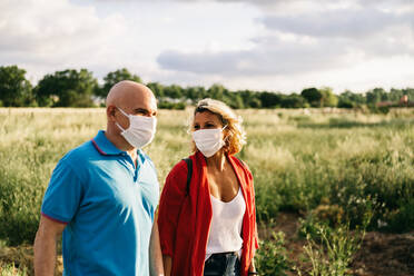Positives erwachsenes Paar mit Gesichtsschutzmasken und Freizeitkleidung, das an einem bewölkten Sommertag an einem ländlichen Feld entlang spaziert - ADSF15609
