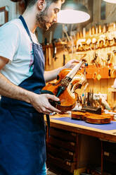 Side view of young craftsman in apron standing with shiny violin near workbench in modern workshop - ADSF15614