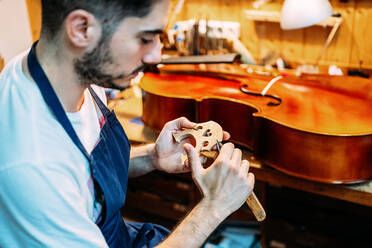 Seitenansicht eines jungen Handwerkers, der mit einem Messer einen hölzernen Geigensteg schnitzt, während er in einer Werkstatt ein Streichinstrument herstellt - ADSF15618