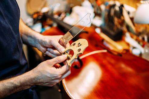 Seitenansicht eines anonymen männlichen Meisters, der einen Geigensteg und professionelles Werkzeug beim Bau eines Musikinstruments in einer Werkstatt hält - ADSF15621