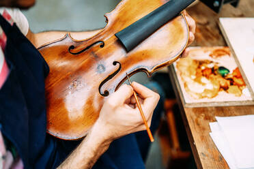 Seitenansicht eines anonymen Handwerkers, der eine antike Geige bei Restaurierungsarbeiten in einer Werkstatt bemalt - ADSF15623