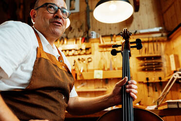 Crop professional senior artisan luthier checking string instrument while working in workshop - ADSF15624