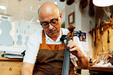 Professional senior artisan luthier checking string instrument while working in workshop - ADSF15625