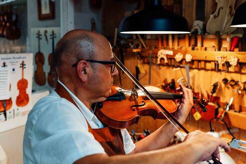 Seitenansicht eines reifen männlichen Geigenbauers, der eine Geige nach der Restaurierung in der Werkstatt prüft - ADSF15627