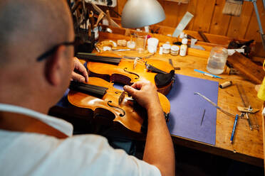 Rückenansicht eines nicht erkennbaren professionellen Geigenbauers, der am Arbeitsplatz sitzt und den Geigensteg auf das Instrument setzt - ADSF15629