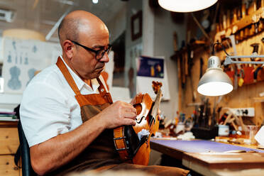 Seitenansicht eines reifen männlichen Geigenbauers mit Schürze und Brille, der auf einem Stuhl sitzt und eine restaurierte Geige hält, während er in der Werkstatt arbeitet - ADSF15630
