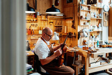 Seitenansicht eines reifen männlichen Geigenbauers mit Schürze und Brille, der auf einem Stuhl sitzt und eine restaurierte Geige hält, während er in der Werkstatt arbeitet - ADSF15632