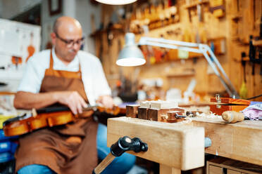 Selektiver Fokus auf ein Detail einer hölzernen Geigenbauer-Werkbank in einer Werkstatt mit einem unscharfen männlichen Handwerker, der eine Geige repariert - ADSF15633