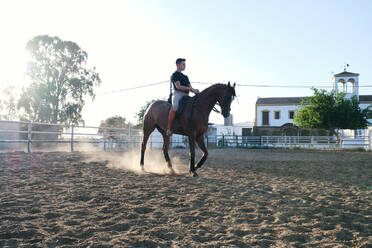 Side view of male equestrian in boots and uniform riding horse on sand arena on ranch during training - ADSF15647