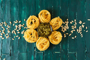 From above of tasty pesto salsa with olive oil surrounded by uncooked pasta rolls and spilled crunchy pine nuts on wooden table - ADSF15658