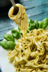 From above of tasty Italian pasta on fork with famous pesto salsa on ceramic plate decorated with fresh basil leaves on wooden table - ADSF15663