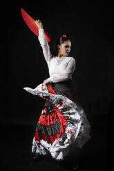 Full body young Hispanic female dancer in colorful costume with red fan in hand performing expressive Flamenco dance on black background - ADSF15668