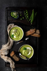 From above view of two bowls of pea cream placed on a wooden tray and dark surface, served with bread and decorated with whole peas and sour cream - ADSF15670