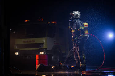 Seitenansicht eines Feuerwehrmanns in Schutzkleidung und Schutzhelm, der in einer regnerischen Nacht in der Nähe eines Feuerwehrautos mit einem Wasserschlauch steht und wegschaut - ADSF15676