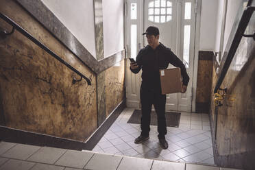 Zusteller benutzt Smartphone, während er ein Paket gegen die Tür hält - MASF19505