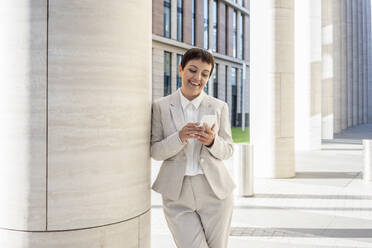 Smiling woman using mobile phone while leaning on pillar at city - VPIF02985