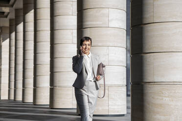 Businesswoman talking on mobile phone while walking against pillar - VPIF02996