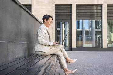 Woman using laptop while sitting on bench against building - VPIF03001
