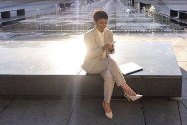 Businesswoman using mobile phone while sitting against fountain at city - VPIF03003