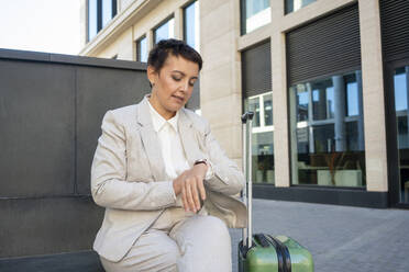 Geschäftsfrau mit Koffer prüft die Zeit, während sie auf einer Bank vor einem Gebäude sitzt - VPIF03011