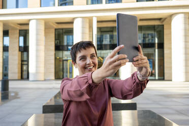 Smiling woman taking selfie with digital tablet while sitting against building at city - VPIF03024