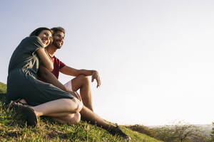Frau umarmt Mann im Gras sitzend gegen klaren Himmel - ABZF03318