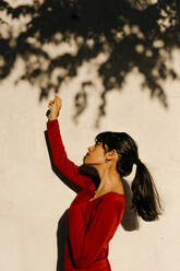 Beautiful woman with hand raised standing by tree shadow on wall - TCEF01043