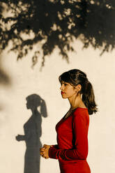 Woman with joined hands standing against wall during sunset - TCEF01048