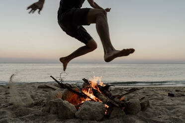 Mann springt bei Sonnenuntergang über Lagerfeuer am Strand - AFVF07232
