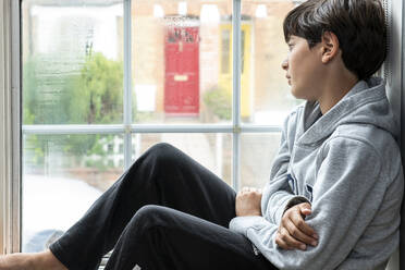 Boy sitting in his bedroom, looking through window during Coronavirus crises. - CUF56477