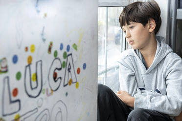 Ein Junge sitzt in seinem Zimmer und schaut während der Coronavirus-Krise durch das Fenster. - CUF56478