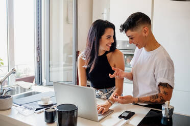 Young lesbian couple standing in kitchen, using laptop. - CUF56480