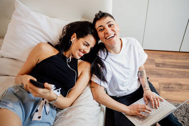 Young lesbian couple in a bedroom, holding mobile phone and laptop, smiling at camera. - CUF56491