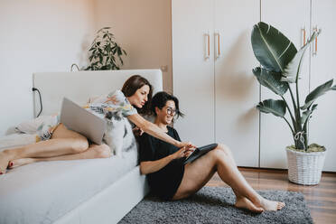 Two women with brown hair sitting on floor and lying on daybed, using laptop and digital tablet. - CUF56522