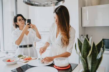 Two smiling women with brown hair standing in a kitchen, preparing food, taking picture with mobile phone. - CUF56536