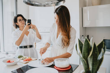 Zwei lächelnde Frauen mit braunen Haaren stehen in einer Küche, bereiten Essen zu und fotografieren mit dem Handy. - CUF56536