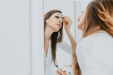 Woman with brown hair standing in front of mirror, applying mascara. - CUF56548