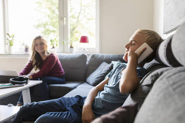 Male teenager talking on phone while smiling sister sitting on sofa at home - MASF19703