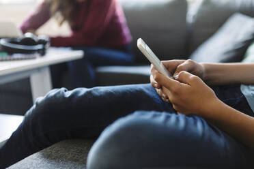 Midsection of teenager using phone while sister sitting on sofa at home - MASF19704