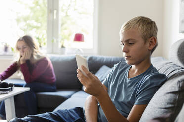 Male teenager using phone while sister sitting on sofa at home - MASF19709