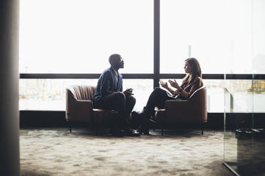 Male and female coworkers discussing while sitting against window at workplace - MASF19734