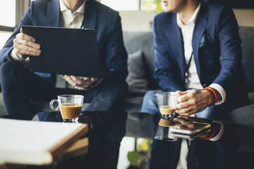 Midsection of businessman showing laptop to colleague while sitting on sofa at workplace - MASF19744