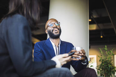 Lächelnder männlicher Unternehmer, der wegschaut, während er mit einer Mitarbeiterin im Büro sitzt - MASF19813