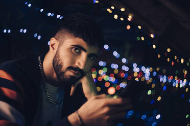 Porträt eines jungen Mannes, der sein Smartphone gegen bunte Lichter in einer Bar hält - MASF19926