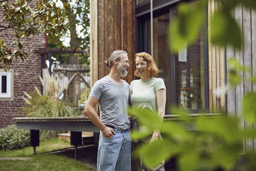 Cheerful couple looking at each other while standing in front of tiny house - MCF01293