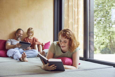 Frau, die ein Buch liest, während Vater und Tochter auf dem Bett zu Hause ein digitales Tablet benutzen - MCF01343