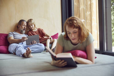 Vater und Tochter benutzen ein digitales Tablet, während die Frau auf dem Bett zu Hause ein Buch liest - MCF01344