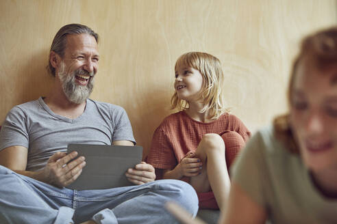 Cheerful father and daughter sitting against wall at home - MCF01346