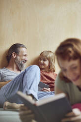 Lächelnder Vater und Tochter schauen sich an, während die Frau zu Hause auf dem Bett ein Buch liest - MCF01349