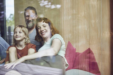 Lächelnde Eltern mit Tochter, die sich zu Hause auf dem Bett ausruhen, durch ein Fenster gesehen - MCF01358